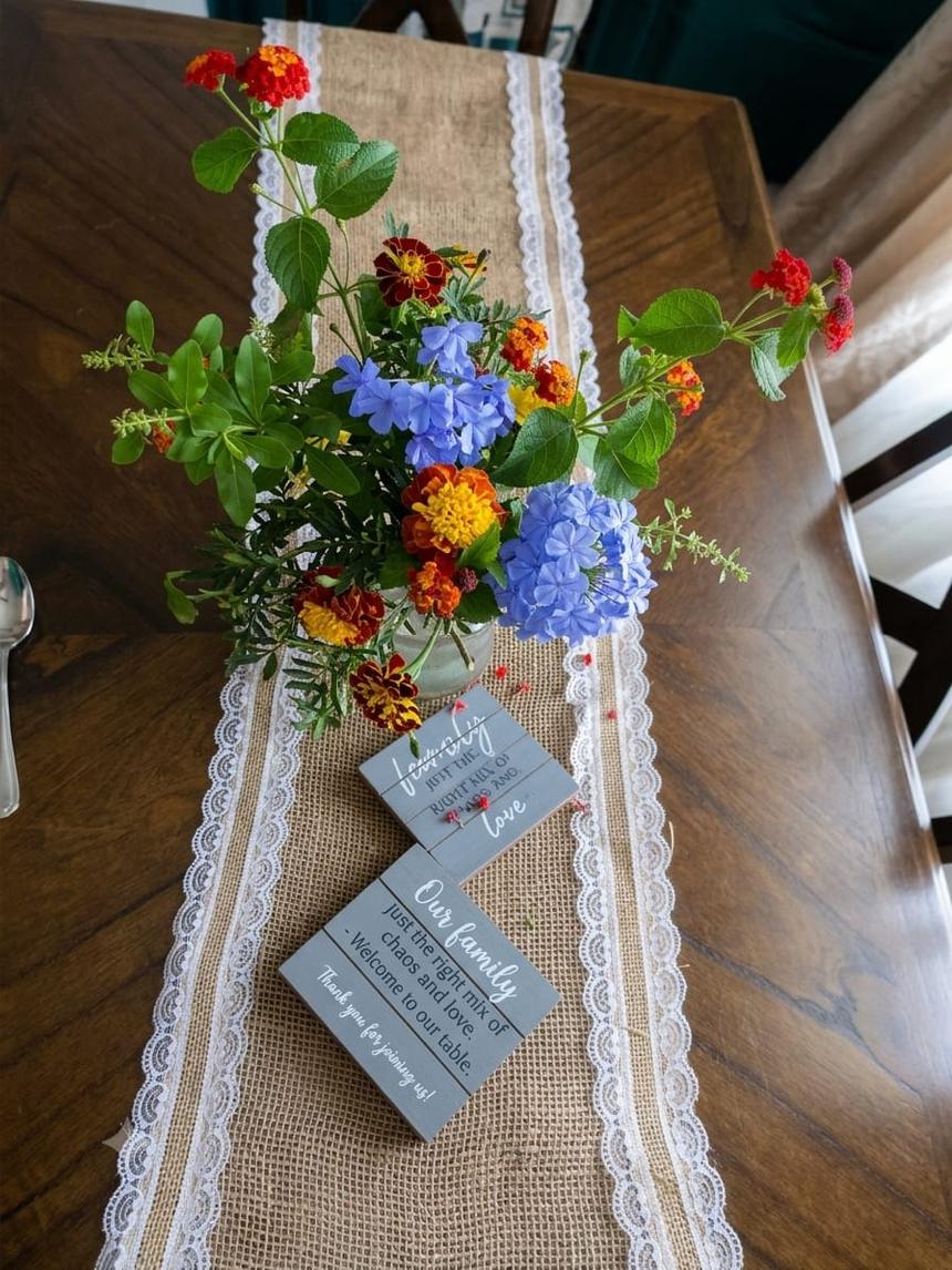 A jar of garden flowers on a burlap runner, beside a hand-painted sign reading: our family, just the right mix of chaos and love. Welcome to our table.